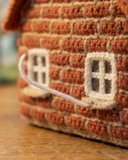 A muted loop of a crochet house front with old windows, where a thin white strand of yarn curls out at a gap