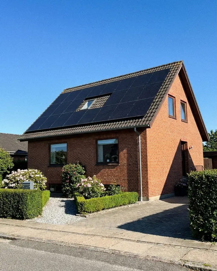A red brick house with a black tiled roof and black solar panels under a clear blue sky, seen from the pavement