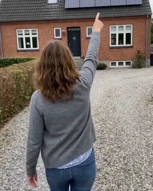 A muted loop of an AI-generated woman, seen from behind, walking up a gravel drive and pointing at the roof of a brick house